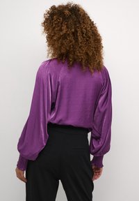 Blusa in satin viola con maniche a palloncino e polsini aderenti, abbinata a pantaloni neri a vita alta, che evidenziano texture e caduta.