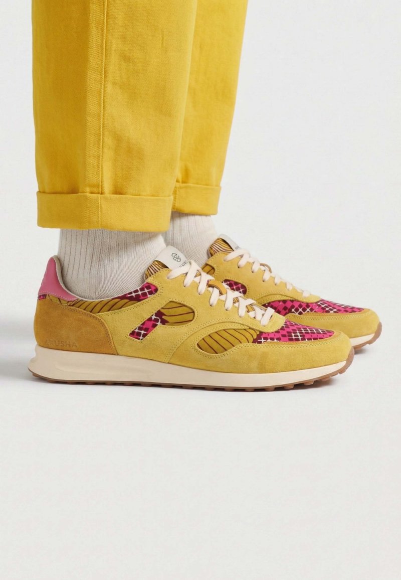 Baskets jaunes et roses à motifs, portées avec des chaussettes blanches et un pantalon jaune retroussé sur fond blanc.