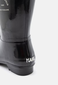 Bota de chuva de borracha preta brilhante com salto baixo, superfície texturizada e um logotipo branco na parte de trás com a inscrição "KARL".