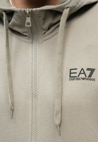 Közelkép egy bézs színű cipzáros kapucnis pulóverről, rajta húzózsinórokkal és fekete EA7 Emporio Armani logóval a mellrészen.