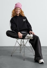 Felpa nera con cappuccio e logo bianco, jeans grigi larghi e scarpe da ginnastica bianche. Il modello è seduto con le gambe incrociate su una sedia di metallo, indossando un berretto rosa.