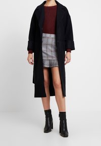 Un long manteau noir avec de larges revers, un pull bordeaux et une jupe mini à carreaux. Des bottines noires avec des zips complètent la tenue.