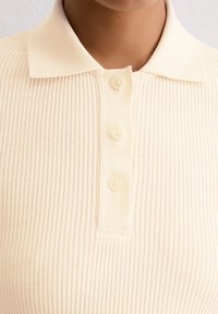 Beige geripptes Poloshirt mit klassischem Kragen, vier Knöpfen und einer weichen Textur. Der Stoff hat ein dezentes vertikales Strukturmuster.