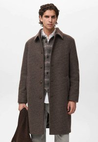 Abrigo de lana marrón con patrón, con un cuello, cierres de botón y bolsillos laterales, superpuesto a una camisa a cuadros gris. El modelo sostiene una chaqueta marrón a juego.