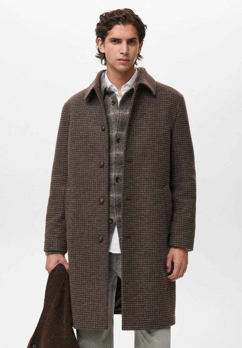 Abrigo de lana marrón con patrón, con un cuello, cierres de botón y bolsillos laterales, superpuesto a una camisa a cuadros gris. El modelo sostiene una chaqueta marrón a juego.