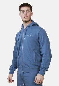 Felpa blu con zip realizzata in tessuto morbido, dotata di cappuccio, tasche frontali e un piccolo logo sul petto. Maniche e fondo fitted.