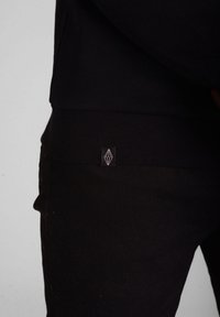 Sweatshirt noir avec une texture douce, arborant un patch de marque en blanc contrasté sur l'ourlet inférieur. Design simple, silhouette ajustée.