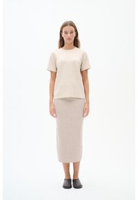 Beige pletené tričko s krátkým rukávem, spárované s odpovídajícími ribbovanými midi sukněmi. Modelka má na sobě černé slip-on boty a stojí na jednolitém pozadí.