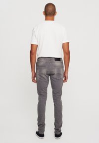 Jeans de denim gris con un ajuste slim, que presentan una etiqueta negra en la cintura trasera y un diseño clásico de cinco bolsillos. Llevados con una camiseta blanca.