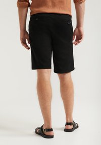 Svarta shorts med rak passform, tillverkade av bomull, med bakfickor och en enkel fåll. Bärs med svarta sandaler.