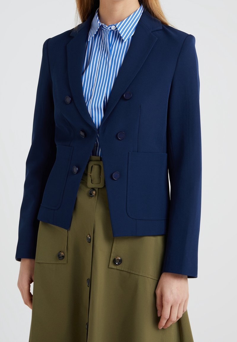 Navy blue double-breasted blazer with round buttons, two front pockets, and structured shoulders, worn over a blue and white striped shirt.