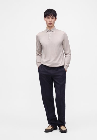 Jeune homme debout les mains dans les poches, portant un polo beige à manches longues, un pantalon noir et des mocassins marron bicolores.