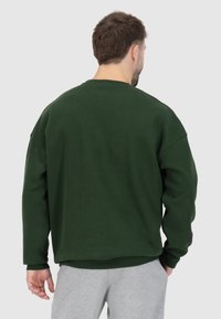 Felpa verde scuro con vestibilità rilassata, spalle cadenti e polsini a coste. Textura morbida, indossata con pantaloni sportivi grigi chiari.