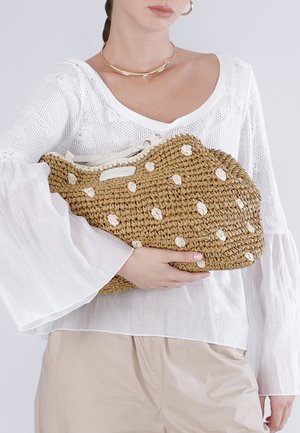 Mujer con blusa blanca y pantalones beige sosteniendo un bolso de mano marrón tejido con detalles de lunares blancos.