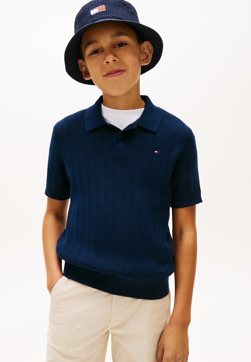 Αγόρι να φοράει μπλε μαρινιέρικη πλεκτή πόλο και καπέλο τύπου κουβά, μπεζ παντελόνι, στέκεται με το ένα χέρι στην τσέπη μπροστά σε λευκό φόντο.