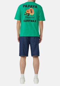 Groene katoenen T-shirt met een kleurrafische papaya-afbeelding en zwarte tekst "PAPAYA geurende SURFWAX" op de achterkant. Gecombineerd met donkere denimshorts.