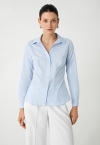 Camisa de botones de manga larga azul claro con collar puntiagudo, diseño ajustado y textura suave. Detalles de botones y puños en color a juego.