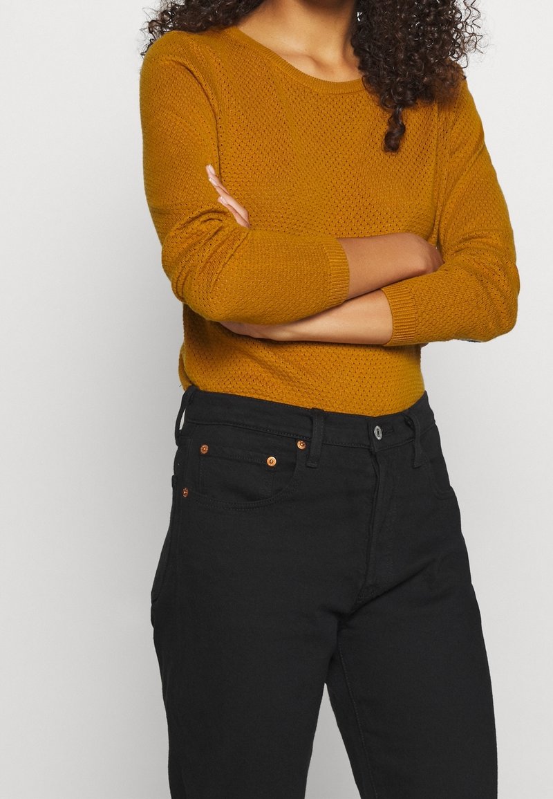 Pull en maille jaune moutarde avec une finition texturée, associé à un jean noir. Le pull a un col rond et des manches trois-quarts.