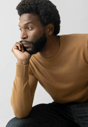Brown long-sleeve knit sweater with ribbed cuffs, worn by a person seated, against a plain white background. Dark jeans visible.