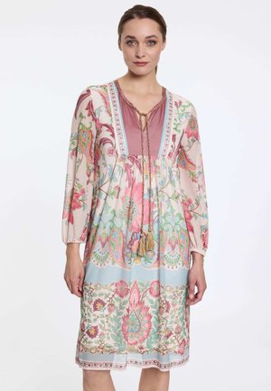 Frau trägt ein knielanges, langärmliges Kleid mit rosa, blauen und grünen Blumenmustern und einer Quastenbinde am Ausschnitt.