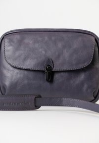 Borsa a tracolla in pelle viola con chiusura a pattina, accessori neri e tracolla rimovibile. Texture liscia con una leggera lucentezza e marchio impresso.