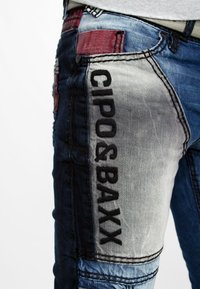 Spijkerbroek met een mix van donkerblauwe en lichtblauwe tinten, geborduurd logo "CIPO & BAXX" aan de zijkant, en contrasterende patchdetails.