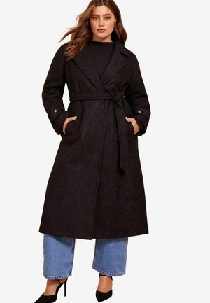 Schwarzer, langer Wollmix-Mantel mit tailliertem Gürtel, großen Revers und Seitentaschen, kombiniert mit weit geschnittenen, verkürzten Jeans und schwarzen Heels.