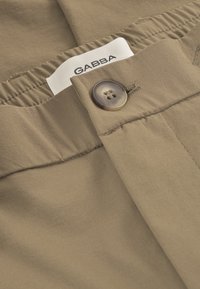 Khaki kalhoty s hladkou texturou, elastickým pasem a kulatým hnědým knoflíkem. Na viditelné bílé etiketě je napsáno "GABBA".