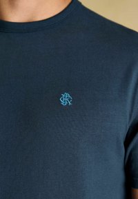 Tuvumā redzams cilvēks, kuram mugurā ir tumši zils krekls ar mazu gaiši zilu izšūtu logotipu uz krūts.