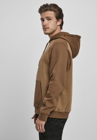 Brun hettegenser med en kontrasterende lysere brun panel, justerbar snor, frontlomme og ribbestrikkede mansjetter og nederkant. Mykt materiale.