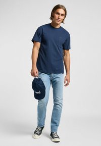 Tummansiniharmaa puuvillainen t-paita lyhyillä hihoilla ja taskulla; yhdistetty vaaleansinisiin farkkuihin ja tummansiniharmaaseen lippalakkkiin, jossa on valkoista tekstiä. Tennarit täydentävät asukokonaisuuden.