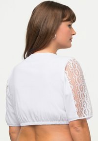 Top bianco corto con maniche corte caratterizzato da accenti in pizzo. Tessuto liscio con orlo arricciato in vita. La vista posteriore mostra capelli e orecchini.
