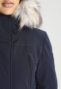 Marinblå parka med en huva i päls, med dragkedja och sidofickor. Materialet verkar slätt med en matt yta.