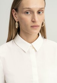 Camisa blanca de cuello con tejido suave, con un cierre frontal abotonado. El sujeto lleva pendientes de aro dorados texturizados. Diseño sencillo.