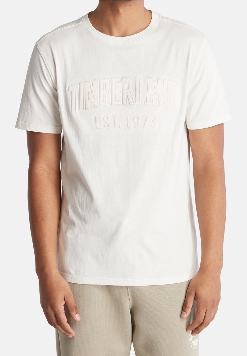 T-shirt blanc en coton avec les inscriptions "TIMBERLAND" et "EST. 1973" en lettres embossées. Manches courtes, col rond, et texture douce.