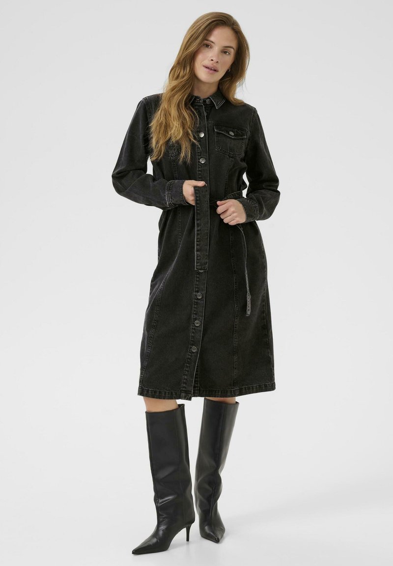 Robe en denim noir à manches longues, avec un bouton à l'avant, deux poches poitrine, une taille cintrée et un ourlet au genou. Assortie à des bottes noires jusqu'au genou.
