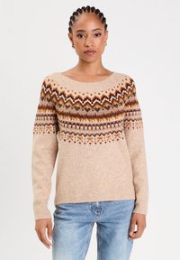 Woman with braided hair wearing gold hoop earrings, a beige patterned knit sweater, and light blue jeans against a plain background.