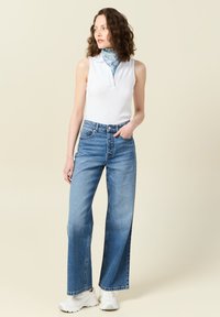 Biała bezrękawnik z dekoltem w kształcie litery V, wysoka talia niebieskich dżinsów i białe trampki. Niebieski szal w wzory związany na szyi. Gładka tekstura.