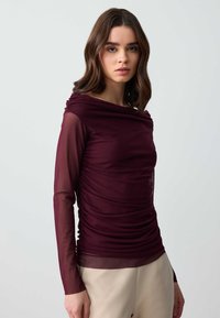 Burgunderfarbener Longsleeve-Oberteil mit geraffter Textur, transparenten Ärmeln und einem drapierten Ausschnitt, kombiniert mit beigen Hosen.