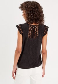 Un haut noir avec un tissu texturé, des manches courtes à volants avec une finition en dentelle, et un panneau en dentelle transparente à l'arrière. Porté avec un pantalon blanc.