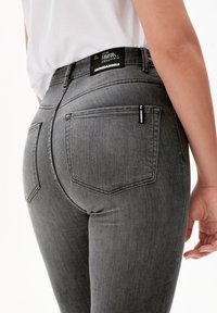 Gros plan d'une personne portant un jean gris taille haute avec des poches arrière, des étiquettes de marque et une chemise blanche rentrée.