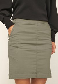 Femme portant une jupe olive longueur genou avec des poches latérales, associée à un sweat à manches longues noir, la main dans la poche.