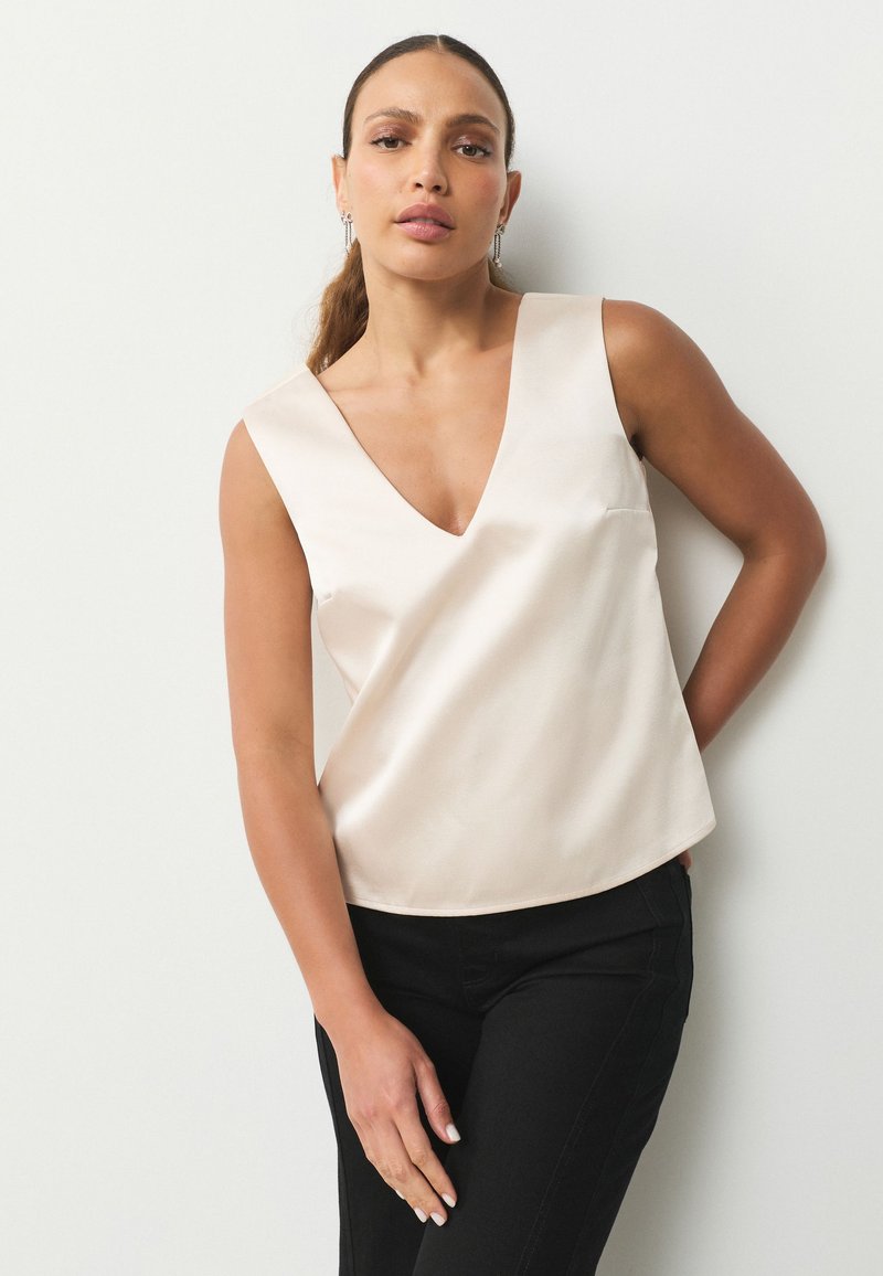 Luźny, bezrękawnikowy top z jasnobeżowego satyny z głębokim dekoltem w serek i subtelnym detalem kieszeni, zestawiony z czarnymi dopasowanymi spodniami.