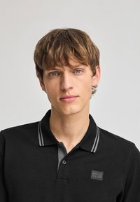 Svart pikétröja med grå randig krage, med en fyrkantig patch med texten "POLO CLUB". Materialet ser slätt ut med en figurnära design.