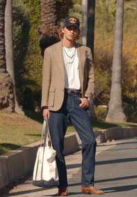 Nuori mies, jolla on päällään beige bleiseri, valkoinen t-paita, tummat farkut, ruskeat saappaat, musta lippalakki, kaulakorut ja hän kantaa valkoista kangaskassia ulkona.