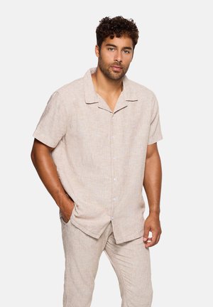 Hombre con cabello rizado, vestido con camisa beige de manga corta abotonada y pantalones a juego, de pie con una mano en el bolsillo frente a un fondo liso.