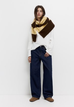Jeans largi din denim albastru, asortați cu un top alb cu mâneci lungi, purtând o eșarfă cu dungi maronii și galbene. Încălțămintea maronie completează look-ul.