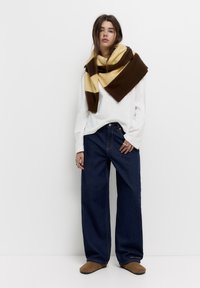 Široké modré džínové kalhoty v kombinaci s bílým topem s dlouhým rukávem, přehnuté hnědým a žlutým pruhovaným šátkem. Hnědá obuv dotváří celkový vzhled.