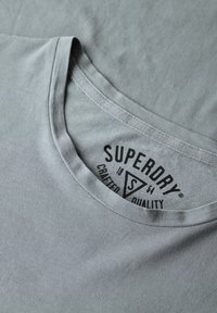 Grijze katoenen t-shirt met een ronde hals. Bevat een zwart logo label binnenin, met de tekst "Superdry, Crafted Quality" en een driehoekontwerp.