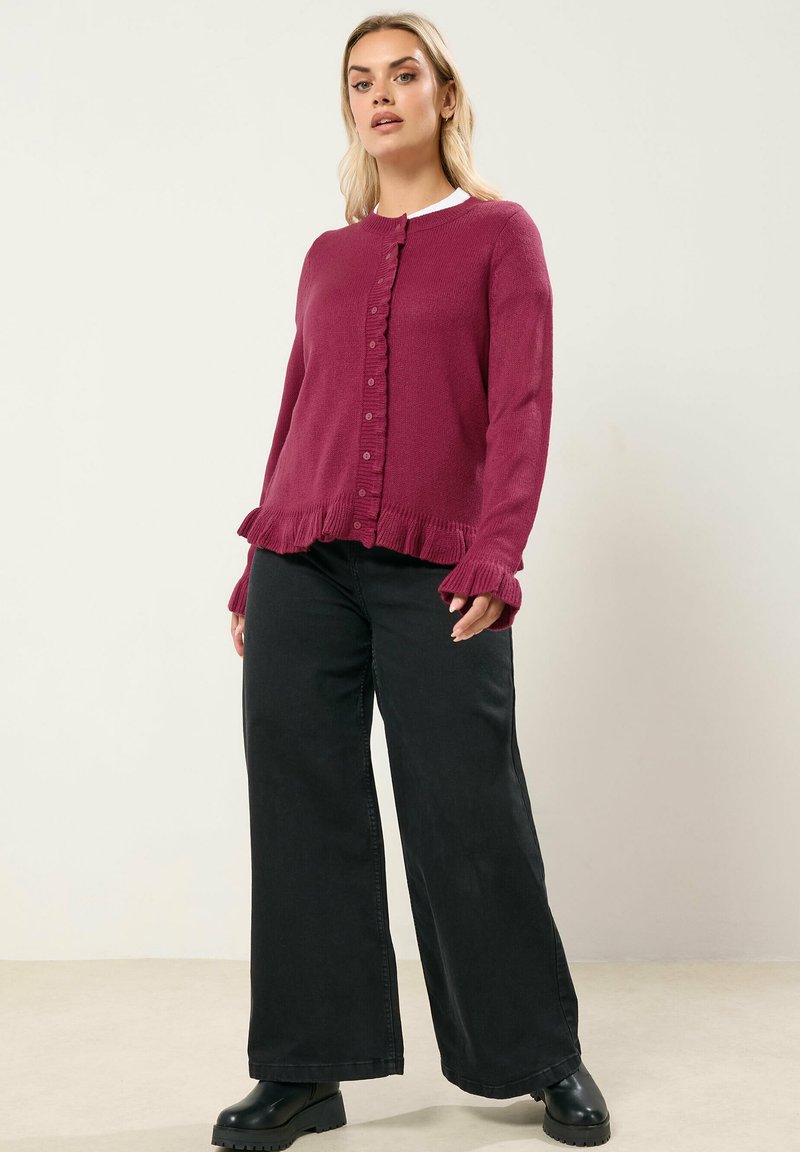 Cardigan en tricot bordeaux avec un devant à boutons, un ourlet et des poignets à volants, associé à un pantalon large noir et des bottes noires épaisses.